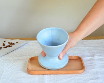 Könnte beinhalten: Hellblaue Keramik-Kaffeegießkanne mit ausgestellter Oberseite und rundem Boden. Sie steht auf einem kleinen, rechteckigen Holztablett. Die Kaffeekanne wird von einer Hand gehalten. Kaffeebohnen sind auf einem weißen Tuch verstreut.