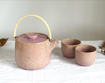 May include: A ceramic tea set in a speckled pink glaze. The set includes a teapot with a bamboo handle and two matching teacups. The teapot has a lid and a spout. The teacups are small and round.