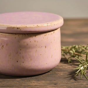 May include: A round, lidded ceramic butter dish in a soft pink hue. The dish has a speckled texture with brown spots and a matching lid. The background is a wooden surface with sprigs of herbs.