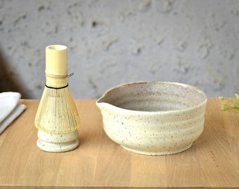 May include: A bamboo matcha whisk and a ceramic matcha bowl with a spout sit on a wooden surface. The whisk is light yellow with a black band. The bowl is speckled with a light beige color.