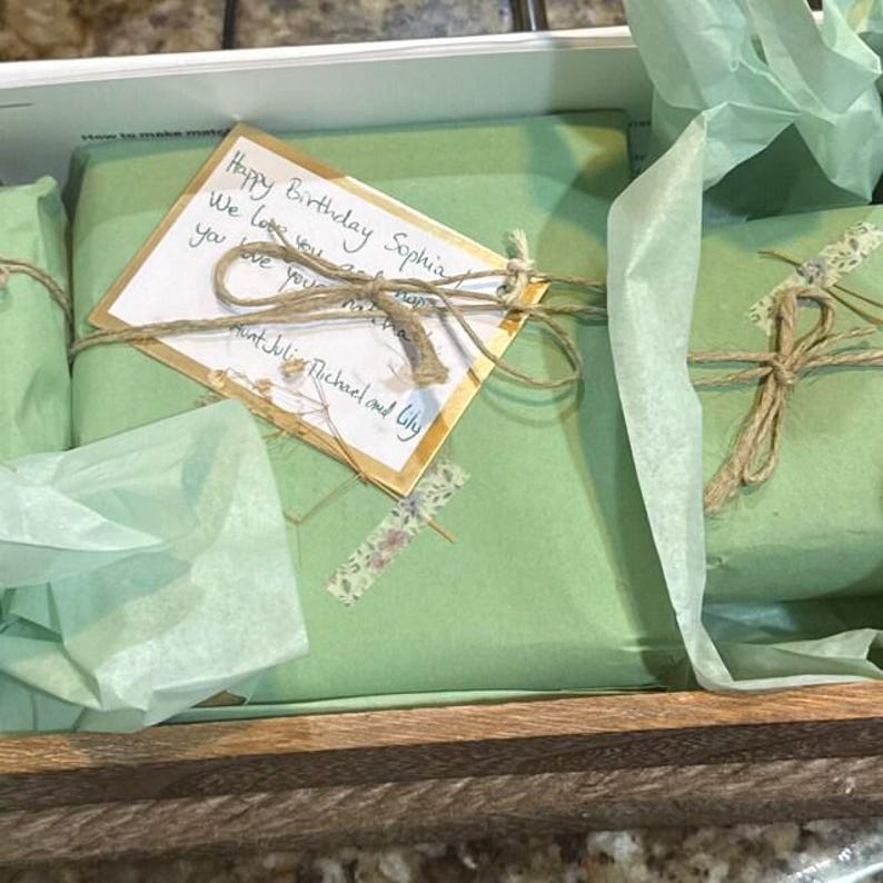 May include: A wooden gift box containing several presents wrapped in light green paper. Each package is tied with twine and has a handwritten card that reads "Happy Birthday Sophia". The gifts are nestled in the box.