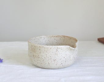 May include: A speckled, off-white ceramic bowl with a pouring spout. The bowl has a rounded shape and is covered in small brown speckles. The bowl is sitting on a white surface.