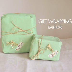 May include: Two gifts wrapped in pale green paper, tied with twine bows. Each package is decorated with dried flowers and a decorative sticker. The text "GIFT WRAPPING available" is visible in the background.