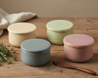 May include: Four small, round ceramic containers in pastel colors: cream, green, blue, and pink. Each container has a matching lid. A wooden butter knife rests on the wooden surface. The containers are likely for storing butter or other spreads.