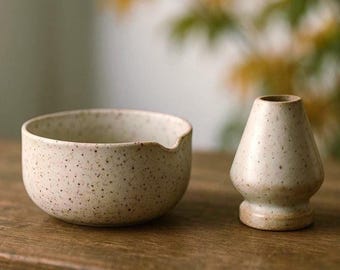 May include: A speckled ceramic matcha bowl with a spout and a matching whisk holder. The off-white pottery has dark brown speckles. The bowl and whisk holder are on a wooden surface.