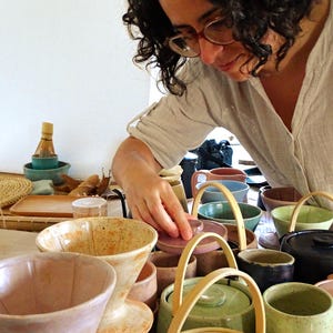 May include: A selection of handcrafted ceramic bowls, cups, and serving dishes in diverse colours, such as pink, green, and blue. Some pieces feature wooden handles or lids. The items are displayed on a table, highlighting the pottery's craftsmanship.