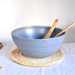 Peut inclure: Un grand saladier en céramique bleu clair avec deux cuillères en bois à l'intérieur, posé sur un dessous de plat en paille tressée. Un distributeur d'huile bleu assorti avec un bec doré est à gauche. Idéal pour les salades.