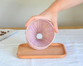 Könnte beinhalten: Ein lavendelfarbener Keramik-Pour-Over-Kaffeebereiter mit einem Holztablett. Der Kaffeebereiter hat eine konische Form mit vertikalen Rippen. Das Holztablett ist rechteckig. Kaffeebohnen sind im Hintergrund zu sehen.