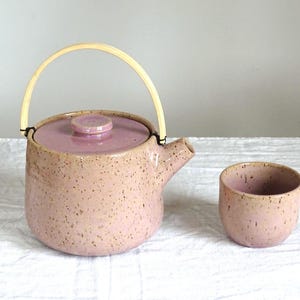 May include: A pink speckled ceramic teapot with a matching lid and a natural bamboo handle. A small, matching cup is next to the teapot. The set is designed for tea service.