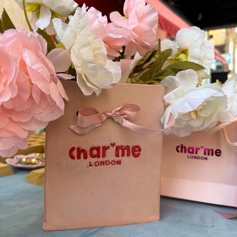 May include: Two pink gift bags with the text "charme LONDON" in red. The bags are tied with a pink ribbon bow and filled with white and pink flowers. The bags are on a blue surface.