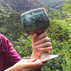 May include: A handcrafted ceramic goblet with a wide bowl and a detailed stem. The bowl features a mountain landscape in shades of green and blue, with the text "The Azzaras" in white. The goblet is held against a backdrop of green foliage and mountains.