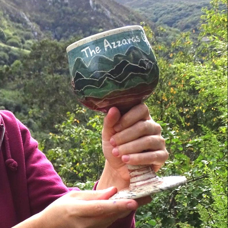 May include: A handcrafted ceramic goblet with a wide bowl and a detailed stem. The bowl features a mountain landscape in shades of green and blue, with the text "The Azzaras" in white. The goblet is held against a backdrop of green foliage and mountains.