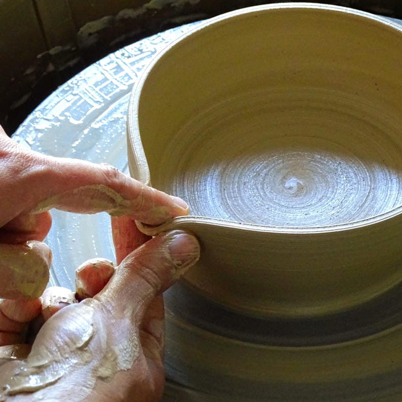 May include: Hands shaping a light brown ceramic bowl on a pottery wheel. The bowl has a wide, shallow design with a curved rim. The hands are covered in wet clay, demonstrating the pottery-making process.