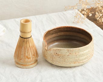 May include: A matcha tea set featuring a ceramic bowl with a spout and a bamboo whisk. The bowl is beige with brown accents, and the whisk is made of light-colored bamboo. The set is designed for preparing matcha tea.