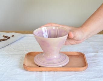 Könnte beinhalten: Ein lila Keramik-Kaffee-Dripper auf einem Holztablett. Der Dripper hat eine konische Form mit vertikalen Rillen und einem kreisförmigen Sockel. Kaffeebohnen sind sichtbar.