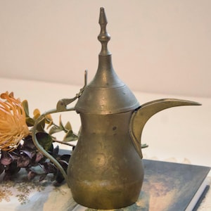May include: A metallic, brass-toned coffee pot featuring a bulbous body, a long, slender, curved spout, and a tall, pointed lid with a finial. The surface shows a weathered, aged patina with subtle, circular etched patterns around the midsection.