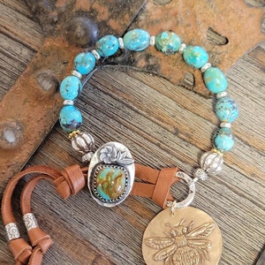 May include: A turquoise and silver beaded bracelet with a leather cord and a silver charm featuring a bee. The bracelet has a turquoise stone set in a silver bezel.