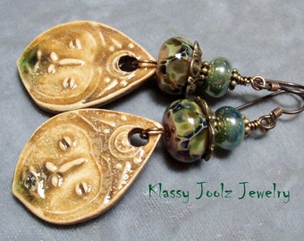 May include: A pair of dangle earrings with ceramic teardrop-shaped charms featuring a face design. The charms are a light brown color with gold accents. The earrings are accented with green and brown glass beads and copper wire.