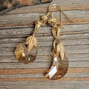 Pode incluir: Brincos dourados com um cristal em forma de lágrima e detalhes de folhas.