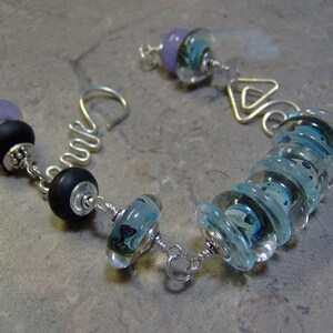 K&ouml;nnte beinhalten: Ein silbernes Armband mit verschiedenen Perlen, darunter ein Stapel blauer Glasperlen, eine schwarze Perle und eine violette Perle.