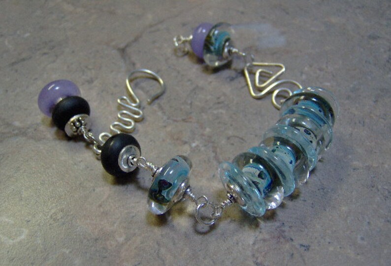 K&ouml;nnte beinhalten: Ein silbernes Armband mit verschiedenen Perlen, darunter ein Stapel blauer Glasperlen, eine schwarze Perle und eine violette Perle.