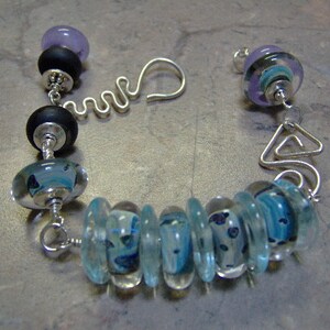 K&ouml;nnte beinhalten: Ein silbernes Armband mit blauen und wei&szlig;en Glasperlen, schwarzen Perlen und lila Perlen.