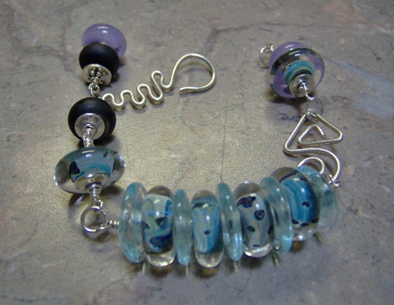 K&ouml;nnte beinhalten: Ein silbernes Armband mit blauen und wei&szlig;en Glasperlen, schwarzen Perlen und lila Perlen.