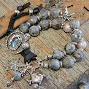 May include: A multi-strand bracelet with a leather cord, silver charms, and gray gemstone beads. The charms include a starfish, a fish, and a round charm with a gemstone in the center. The bracelet is on a wooden surface.