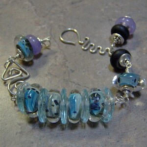 K&ouml;nnte beinhalten: Ein silbernes Armband mit blauen und violetten Glasperlen. Das Armband hat einen silbernen Verschluss und eine silberne Kette.