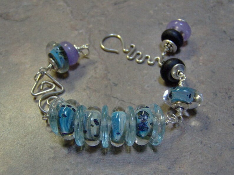 K&ouml;nnte beinhalten: Ein silbernes Armband mit blauen und violetten Glasperlen. Das Armband hat einen silbernen Verschluss und eine silberne Kette.