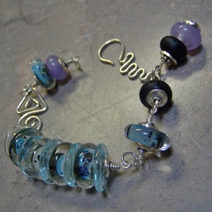 K&ouml;nnte beinhalten: Ein silbernes Armband mit verschiedenen blauen und violetten Glasperlen. Das Armband hat ein einzigartiges Design mit einem Silberdraht, der eine Dreiecksform bildet.