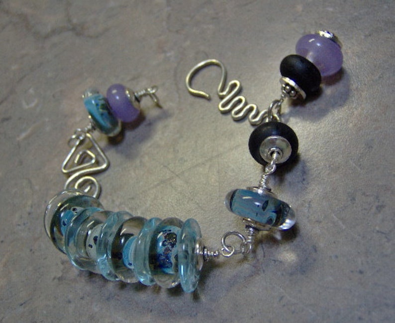 K&ouml;nnte beinhalten: Ein silbernes Armband mit verschiedenen blauen und violetten Glasperlen. Das Armband hat ein einzigartiges Design mit einem Silberdraht, der eine Dreiecksform bildet.