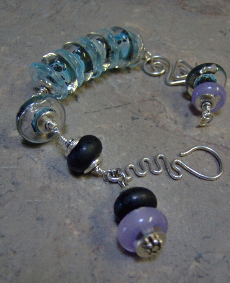 K&ouml;nnte beinhalten: Ein silbernes Armband mit einer Reihe von blauen und wei&szlig;en Glasperlen, zwei schwarzen Perlen und einer lila Perle mit einem silbernen Anh&auml;nger. Das Armband hat einen silbernen Verschluss.