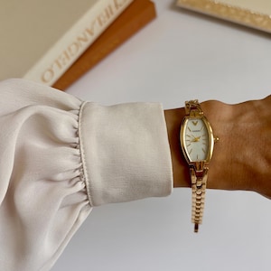 Könnte beinhalten: Goldfarbene Armbanduhr mit ovalem Zifferblatt und weißem Zifferblatt. Die Uhr hat ein Gliederarmband und wird am Handgelenk getragen. Das Zifferblatt der Uhr hat goldene Zeiger und Markierungen. Ein klassisches, elegantes Accessoire.