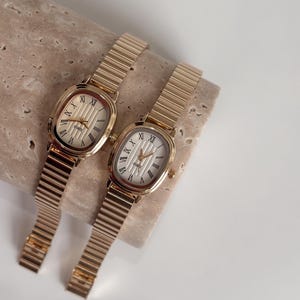 May include: Two gold-toned watches with oval faces and Roman numeral hour markers. The watch faces are white with vertical lines. The watch bands are made of linked metal. The watches are displayed on a light-colored, textured surface.