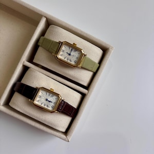 Könnte beinhalten: Zwei rechteckige, goldfarbene Armbanduhren mit römischen Ziffern und blauen Zeigern. Eine Uhr hat ein grünes Lederarmband, die andere ein braunes Lederarmband. Die Uhren werden in einer beigen Box mit gepolsterten Fächern präsentiert.