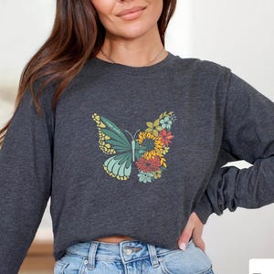 May include: Dark grey heather long-sleeve shirt featuring a butterfly design. The butterfly has a teal and yellow wing with a floral design on the other wing. The text "Dark Grey Heather" is in a white box.