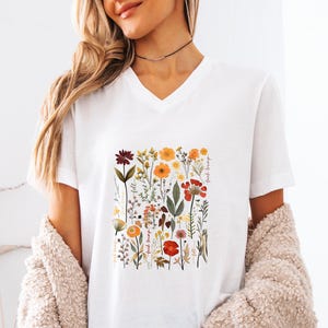 May include: White V-neck t-shirt featuring a colorful floral design with red, orange, and yellow flowers. The words "feel strong" are printed on the right side of the floral design. The shirt is made of a soft material.