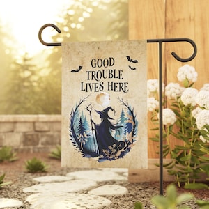 May include: A rectangular garden flag with a beige, parchment-like background. The text "GOOD TROUBLE LIVES HERE" is printed in black above a circular illustration of a witch in a pointed hat holding a staff, surrounded by trees and a moon.