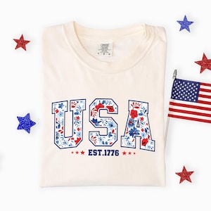 May include: Cream-colored t-shirt with the word "USA" in blue outlined letters filled with a floral pattern of red and blue flowers. The text "EST. 1776" is below the word "USA". The shirt is surrounded by red and blue star-shaped decorations and a small American flag.