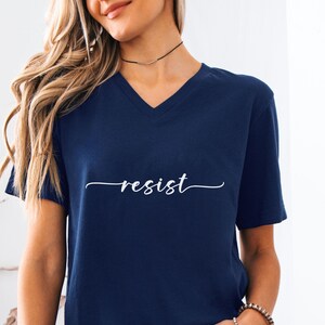 May include: Navy blue V-neck t-shirt with the word "resist" in a white script font. The shirt is a solid color and has short sleeves. The word "Navy" is printed in white in the bottom right corner.