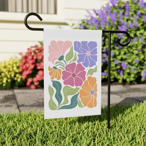 May include: A white garden flag featuring a floral design with pink, orange, purple, and green elements. The flag includes the text "Fuck Trump" in a repeating pattern around the flowers. The flag is displayed on a black metal stand in a grassy yard.