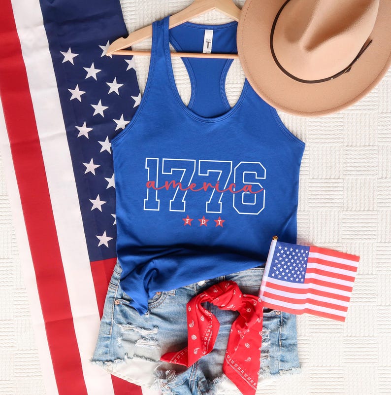 May include: A blue vest top with the text "1776 America" in white and red, alongside denim shorts, a red bandana, and a small American flag. A brown cowboy hat is also present, creating a patriotic theme.