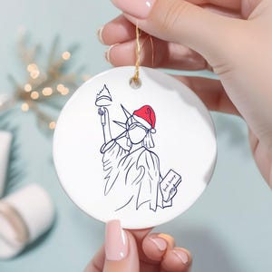 May include: White ceramic Christmas ornament featuring a line drawing of the Statue of Liberty wearing a Santa hat. The statue holds a torch and a tablet with the words "fuck trump". The ornament has a gold string for hanging.