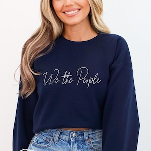 May include: A navy blue sweatshirt featuring the white script text "We the People". The long-sleeved top is made of a soft material and is suitable for casual wear. The sweatshirt is paired with blue denim jeans.