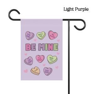 May include: A light purple garden flag with the words "BE MINE" in pink and green. The flag features heart-shaped candies with phrases like "Free Press" and "Civil Rights". The flag is displayed on a black metal stand.