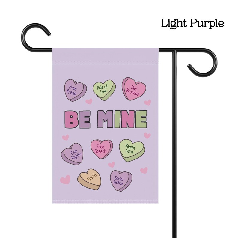 May include: A light purple garden flag with the words "BE MINE" in pink and green. The flag features heart-shaped candies with phrases like "Free Press" and "Civil Rights". The flag is displayed on a black metal stand.