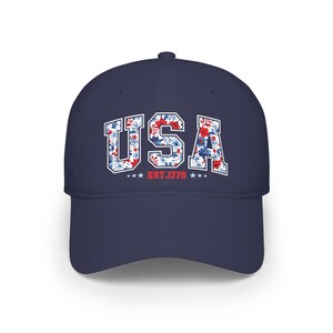 May include: A navy blue baseball cap with the word "USA" in white block letters filled with a floral pattern of red, white, and blue flowers. Below the text are three stars and the text "EST. 1776".