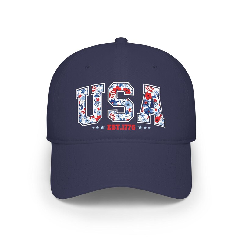 May include: A navy blue baseball cap with the word "USA" in white block letters filled with a floral pattern of red, white, and blue flowers. Below the text are three stars and the text "EST. 1776".