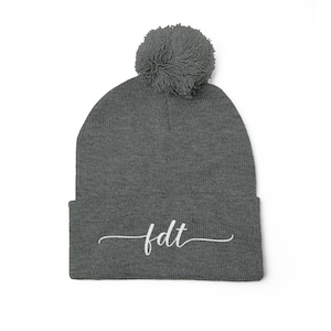May include: A grey beanie hat with a matching pom-pom. The hat has the embroidered letters "fdt" in white, cursive script. The beanie is made of a soft, knitted material.